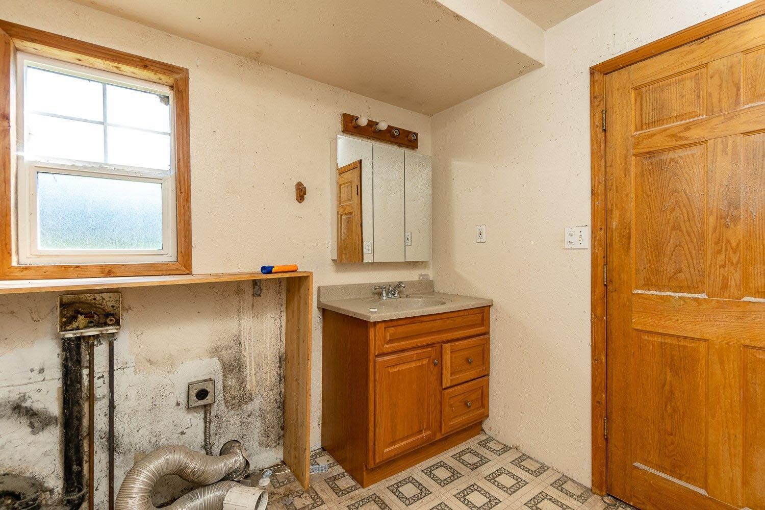 Bathroom with vanity, mirror, and window. Walls show water damage, and a door is visible.