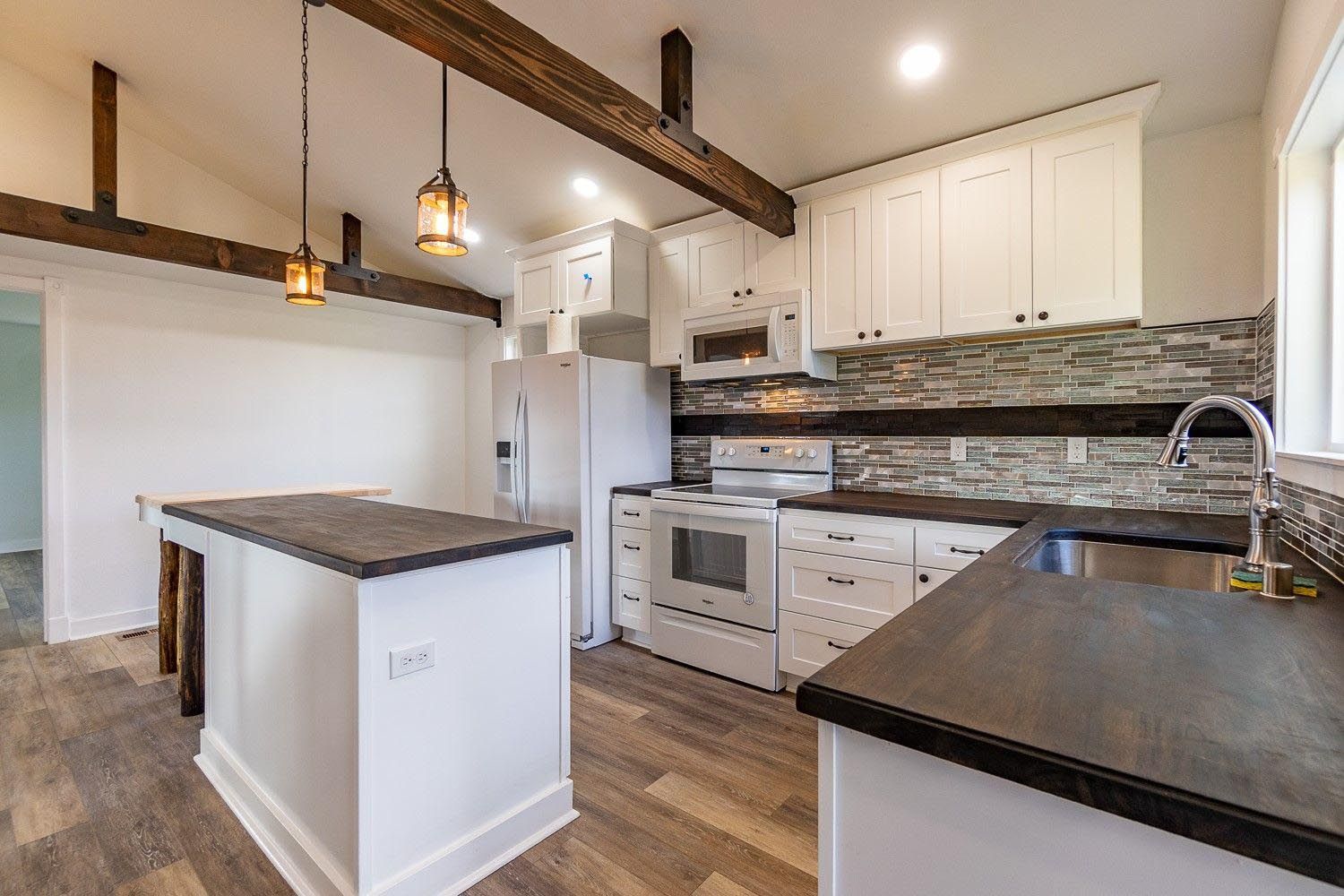 Modern white kitchen with island, wooden beams, white appliances, and a gray backsplash.