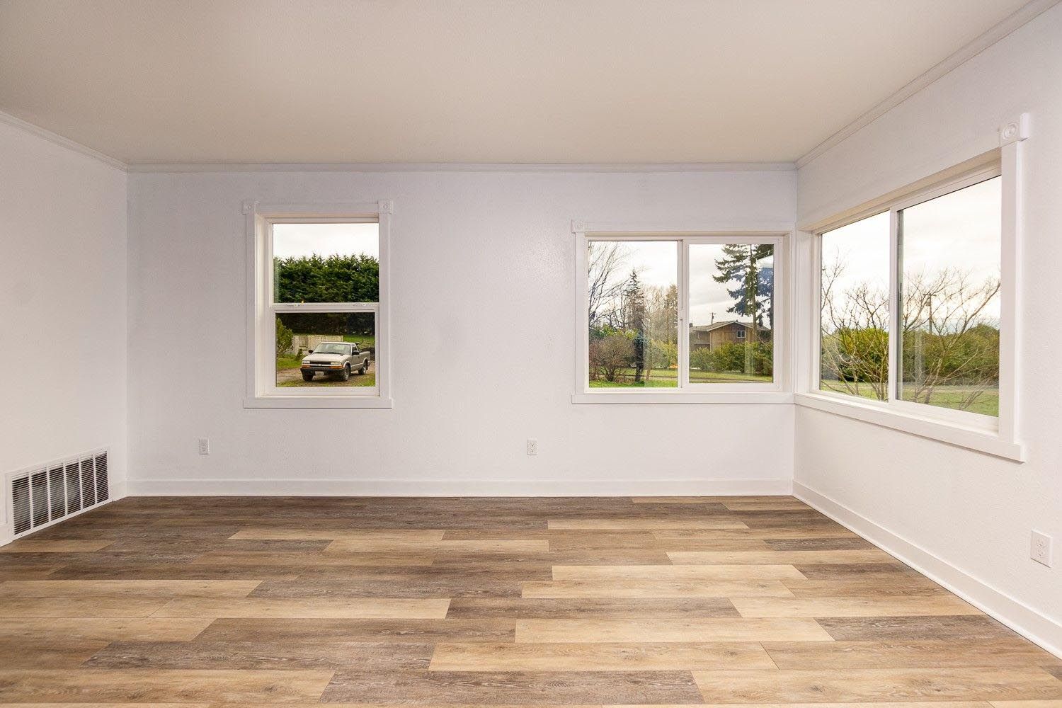 Empty room with wood-look floor and white walls. Windows overlook a yard.