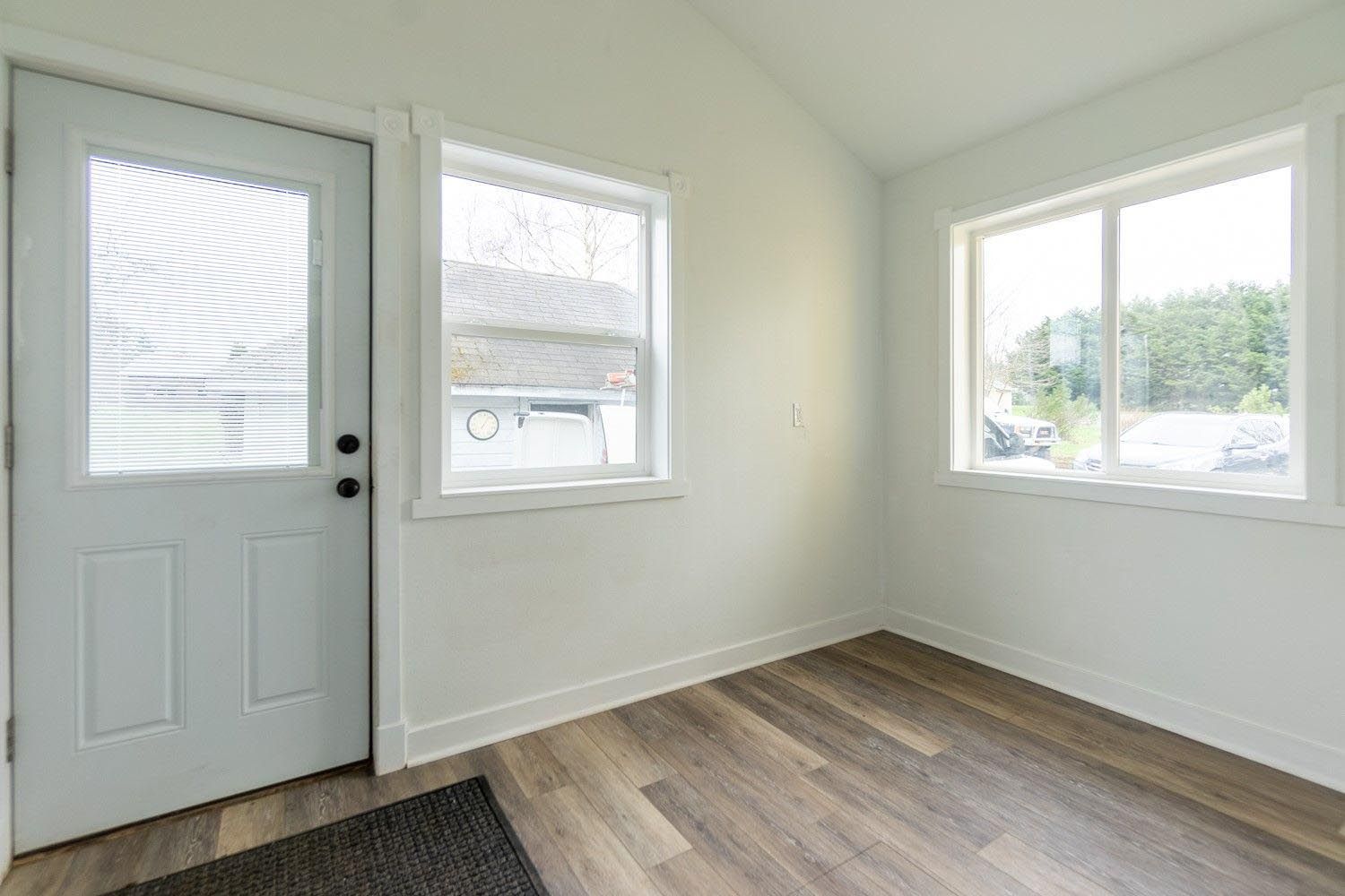 Bright, empty room with a door, two windows, wood-look flooring, and white walls.