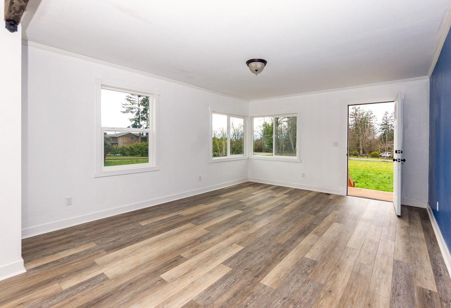 Empty room with light wood-look flooring, white walls, and blue accent wall; three windows and open doorway to a green yard.