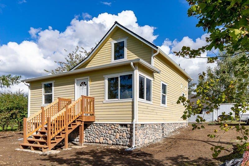 Yellow two-story house with wooden stairs and rock foundation under a bright blue sky.