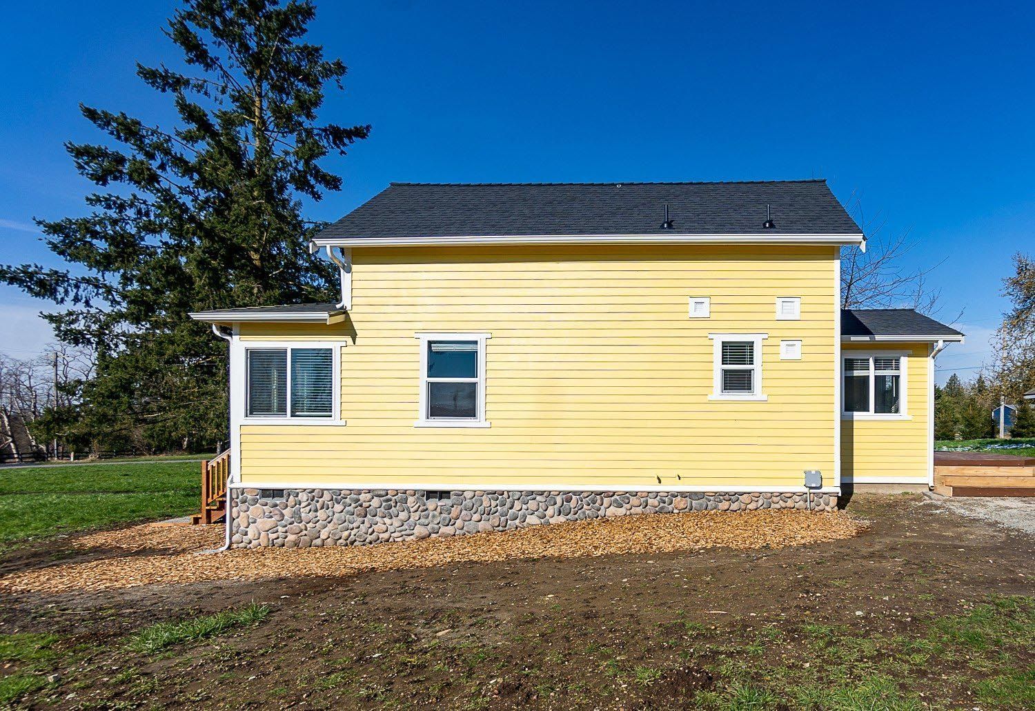 Yellow house with dark roof and rock foundation, set against blue sky.