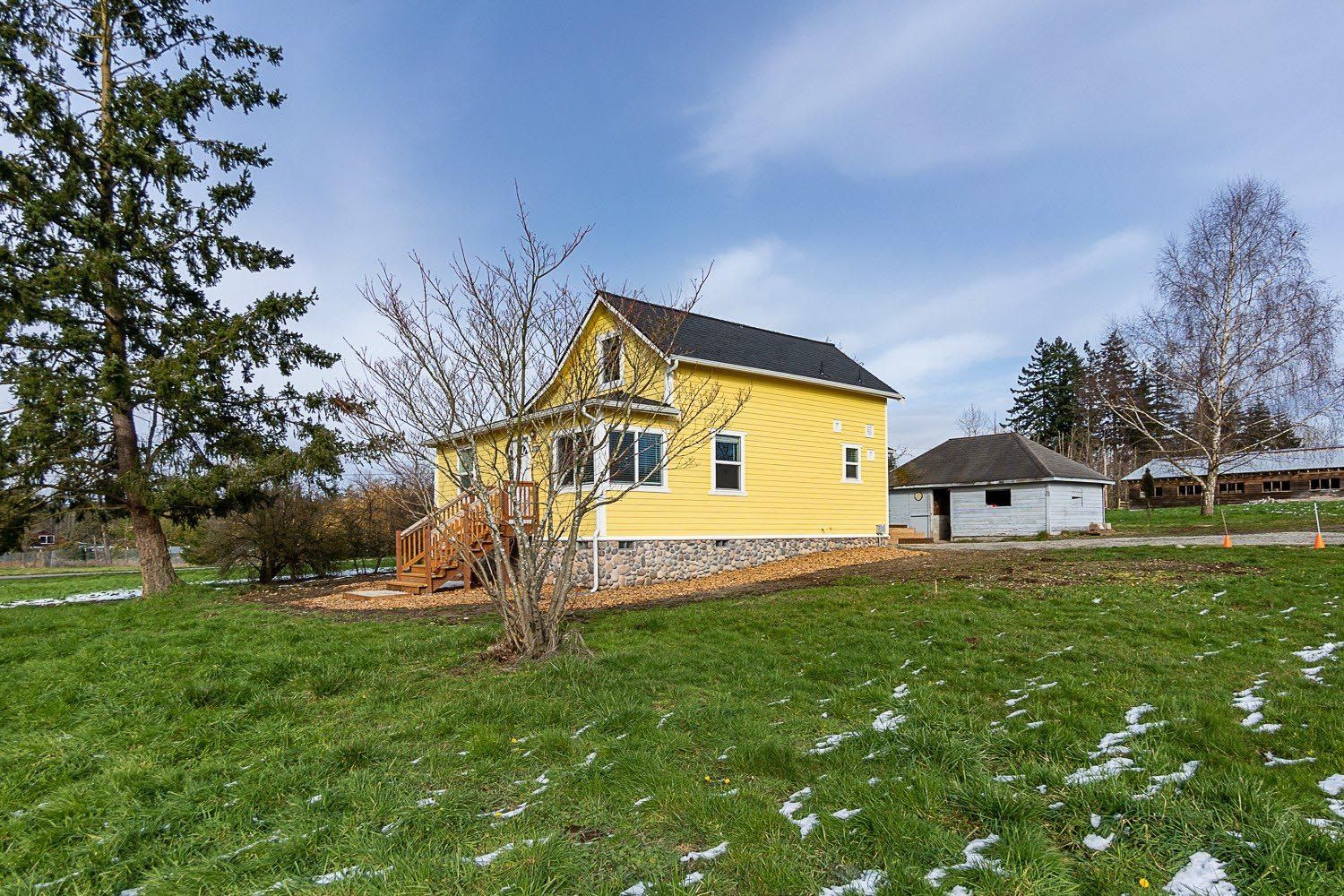 Yellow house with a small porch sits on a grassy lot with patches of snow. A small white building is behind it.