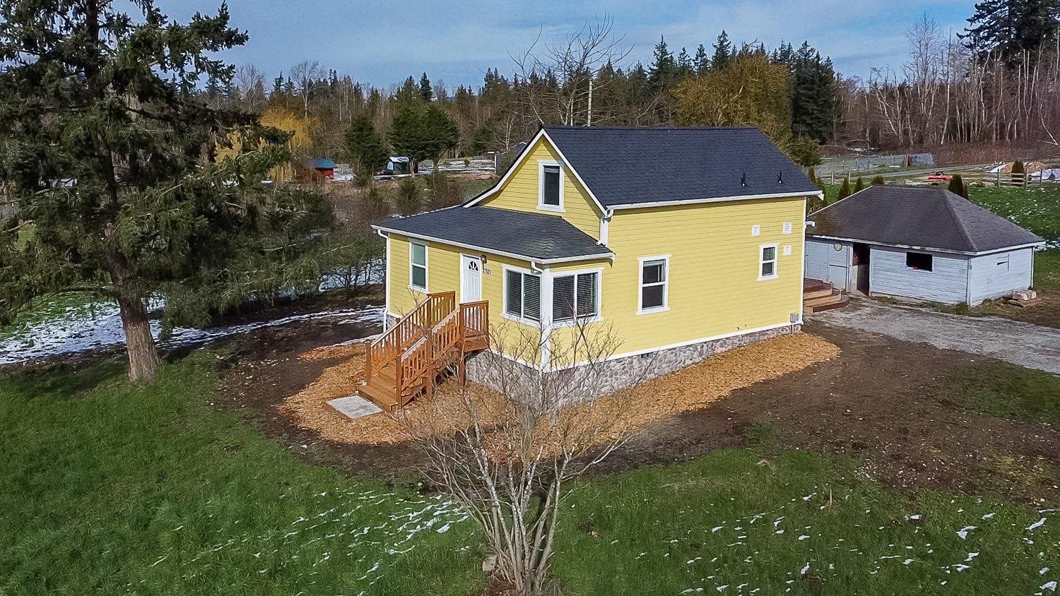 Yellow house with wooden porch, gray roof, and small detached building in a grassy rural setting.