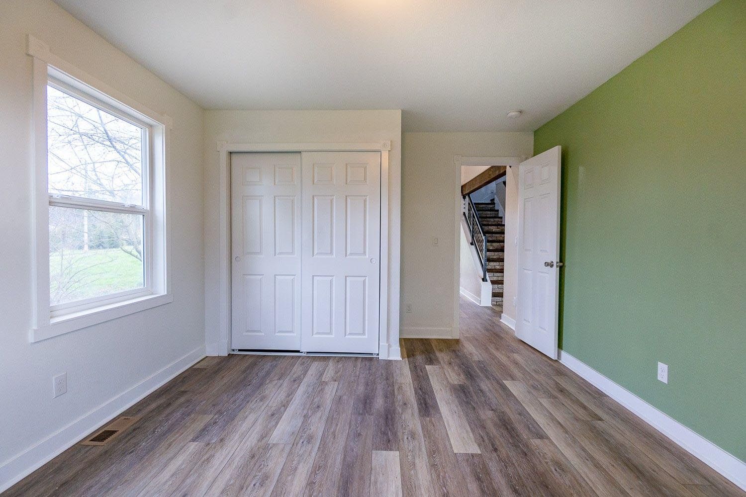Empty room with wood floors, closet, window, and a green accent wall. Doorway leads to stairs.