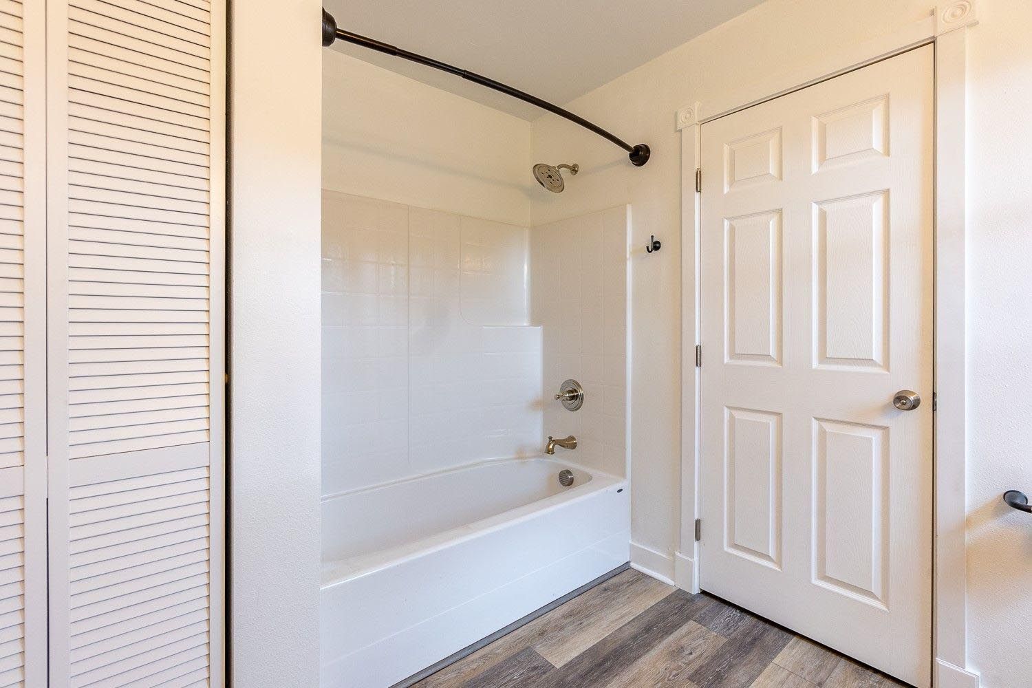 Bathroom with white tub, shower, and door; grey wood-look flooring.