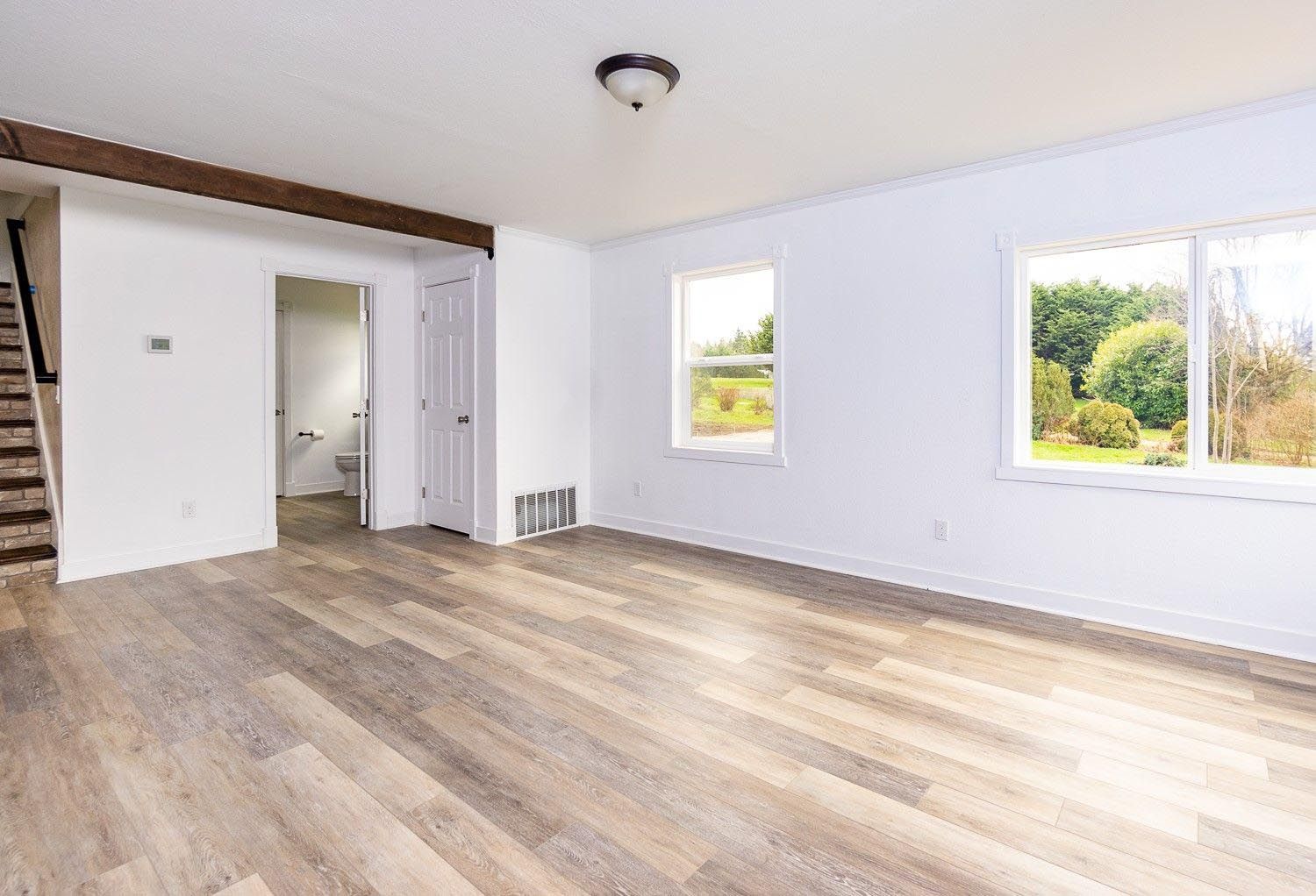 Empty living room with wood-look flooring, white walls, and two windows overlooking a yard.