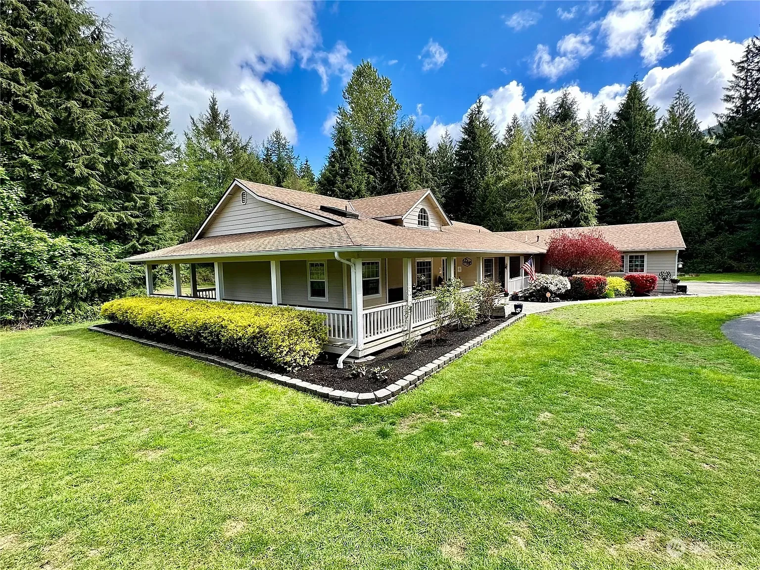 House with wrap-around porch, green lawn, and landscaping, against a backdrop of trees and a partly cloudy sky.