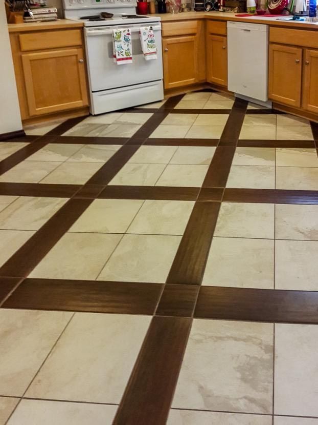 Kitchen floor with cream tiles, framed by dark brown tiles. Cabinets and appliances are visible.