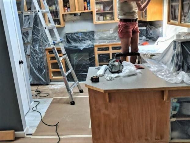Person painting kitchen cabinets, standing on a ladder. Kitchen is covered with plastic sheeting for protection.