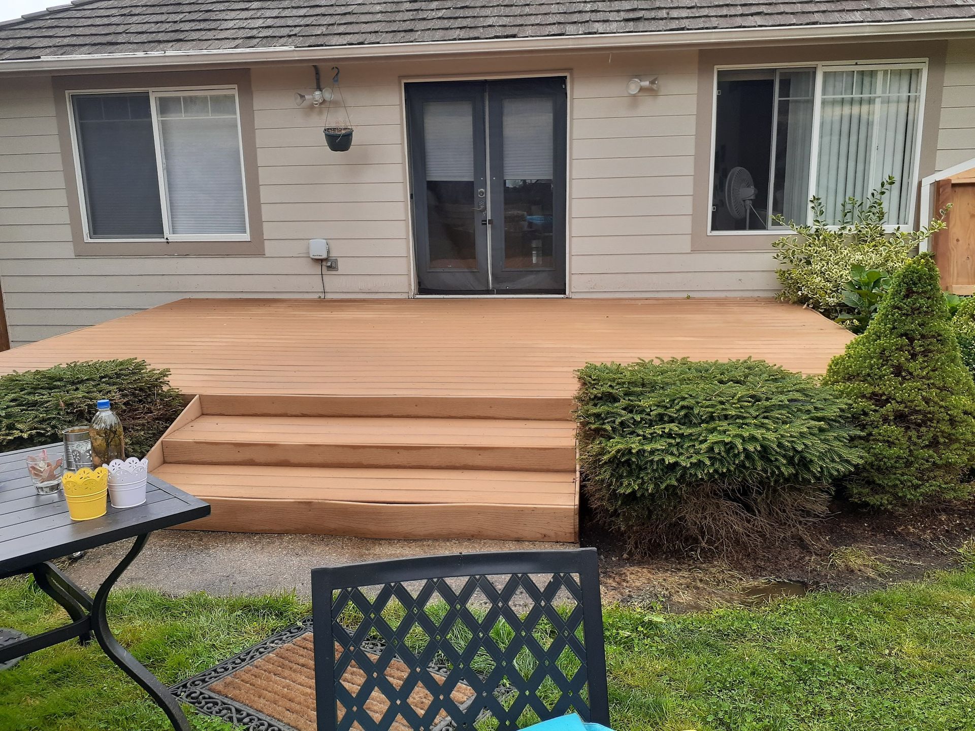 Brown wooden deck with steps, leading to a house with windows and a door, surrounded by greenery.