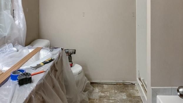 Bathroom renovation in progress; tools on covered toilet, plastic sheeting, tan walls, exposed flooring.