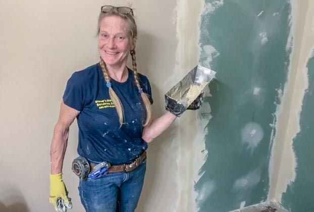 Woman in blue shirt and jeans, smiling, holding a putty knife while repairing drywall.