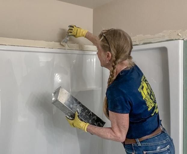 Woman in a blue shirt and jeans using a trowel to apply drywall mud around a shower stall.