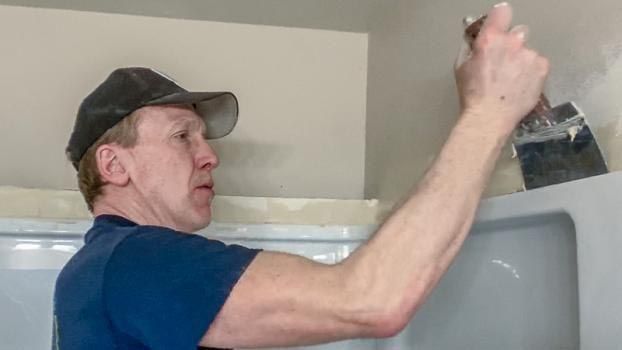 Man in a cap, uses a putty knife, applying something to a light-colored wall, near a white shower.