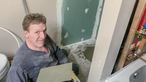 Man installing tile in a bathroom. He’s kneeling, holding tile and trowel, looking at the camera.