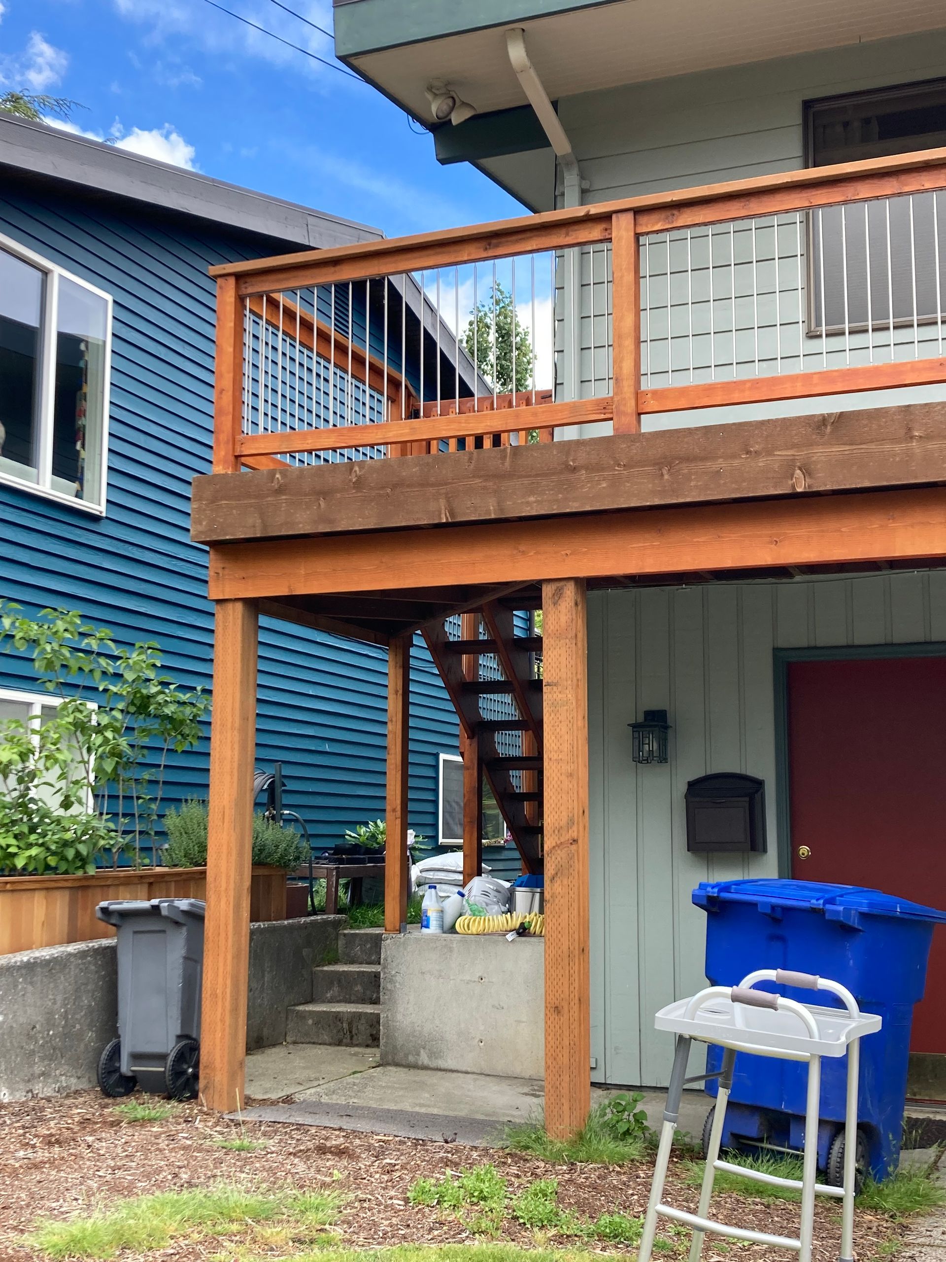 Two-story building exterior. Blue and green siding, wooden deck, stairs, trash bins.
