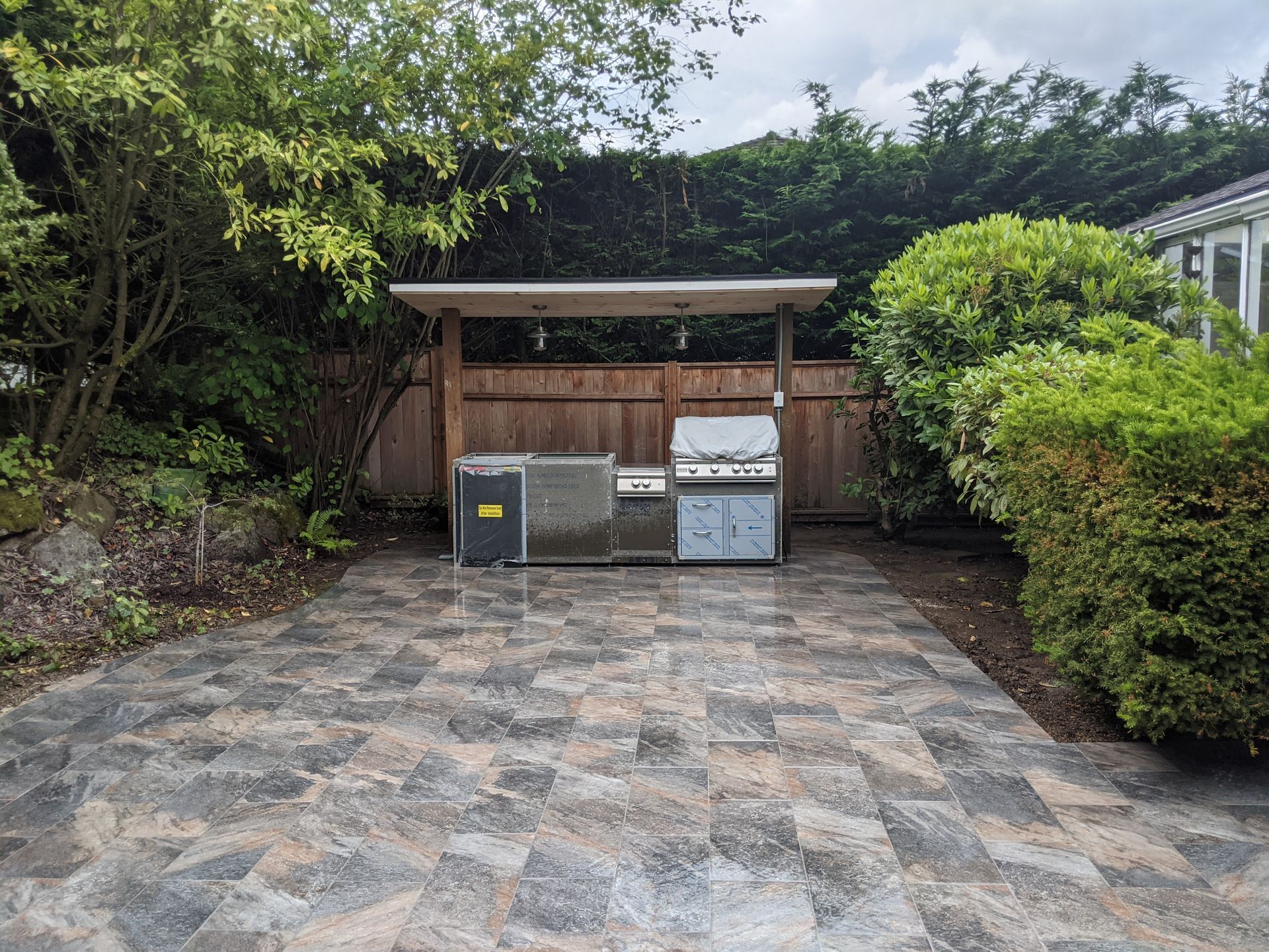 Outdoor kitchen on a paved patio with a grill, counter, and wooden structure.