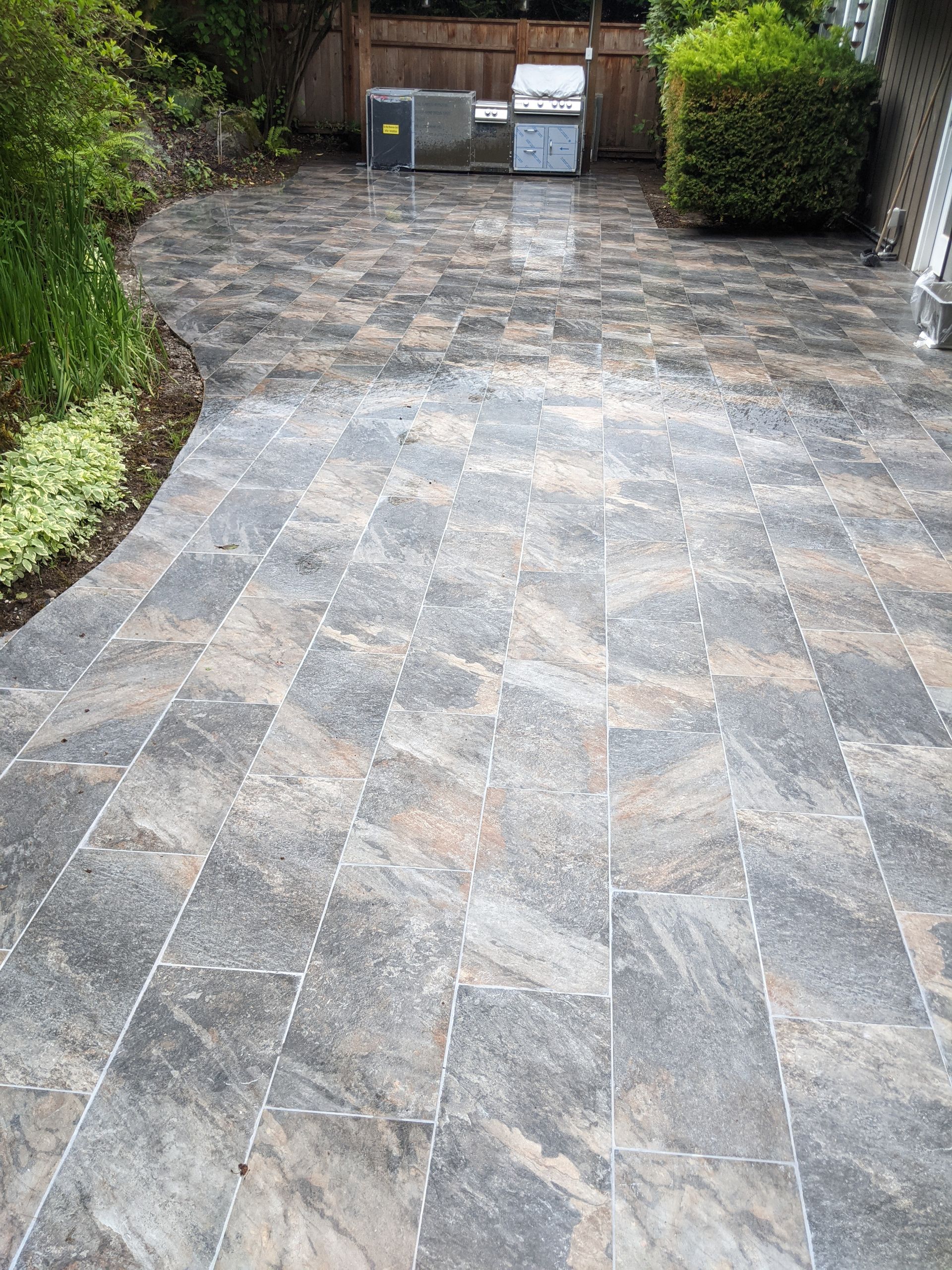 Patio paved with rectangular, marbled gray and tan pavers. Outdoor kitchen in background.