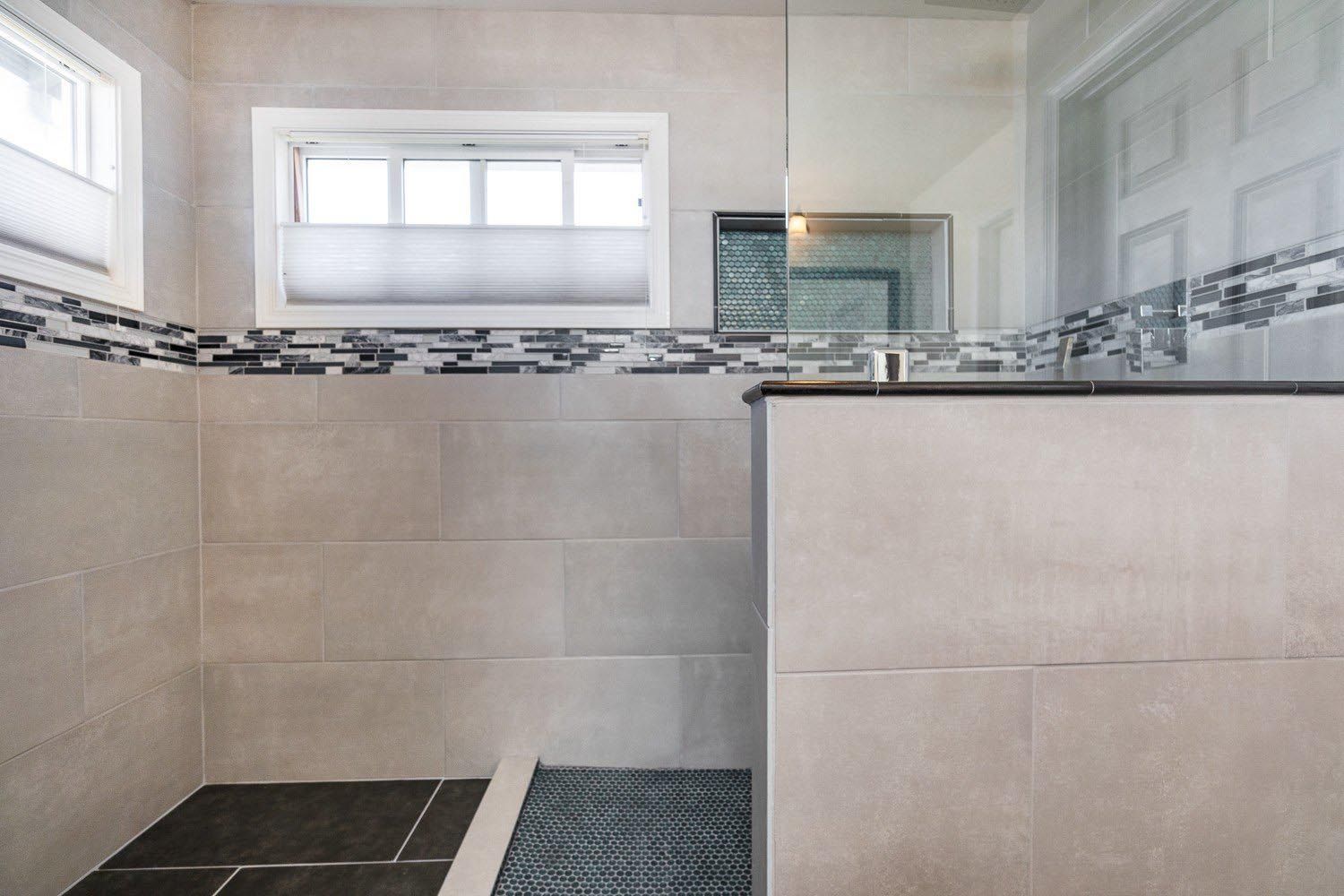 Bathroom with large gray tiles, a glass shower, and a small window with a shade.