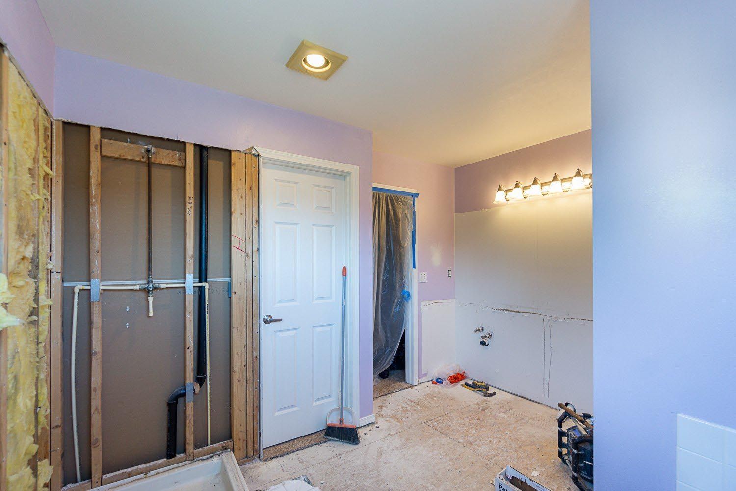 Bathroom renovation in progress, walls open, showing studs, plumbing, and a door, pale purple walls.