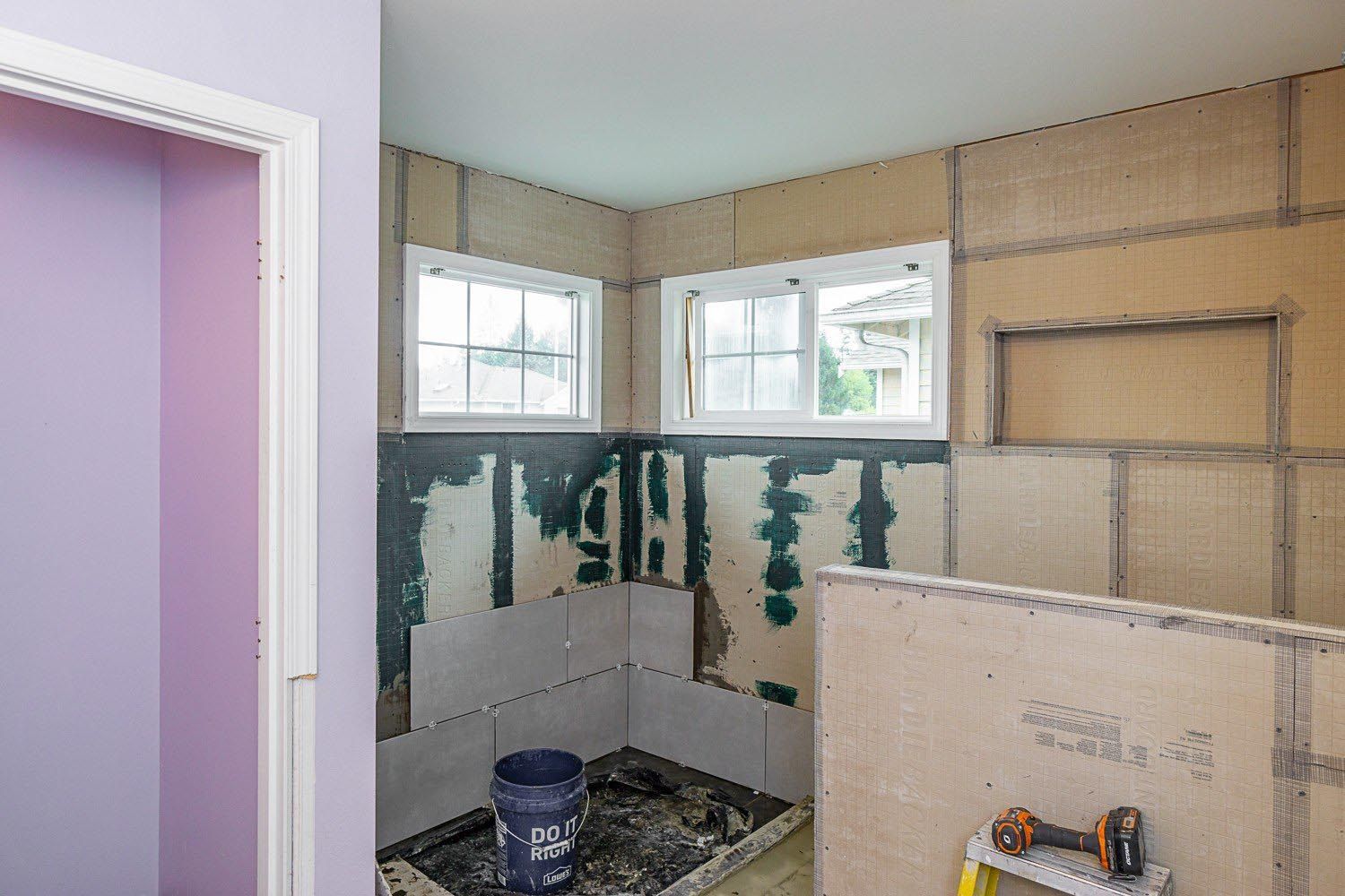 Bathroom renovation in progress, walls with cement board, partially tiled, with bucket, windows, and drywall.