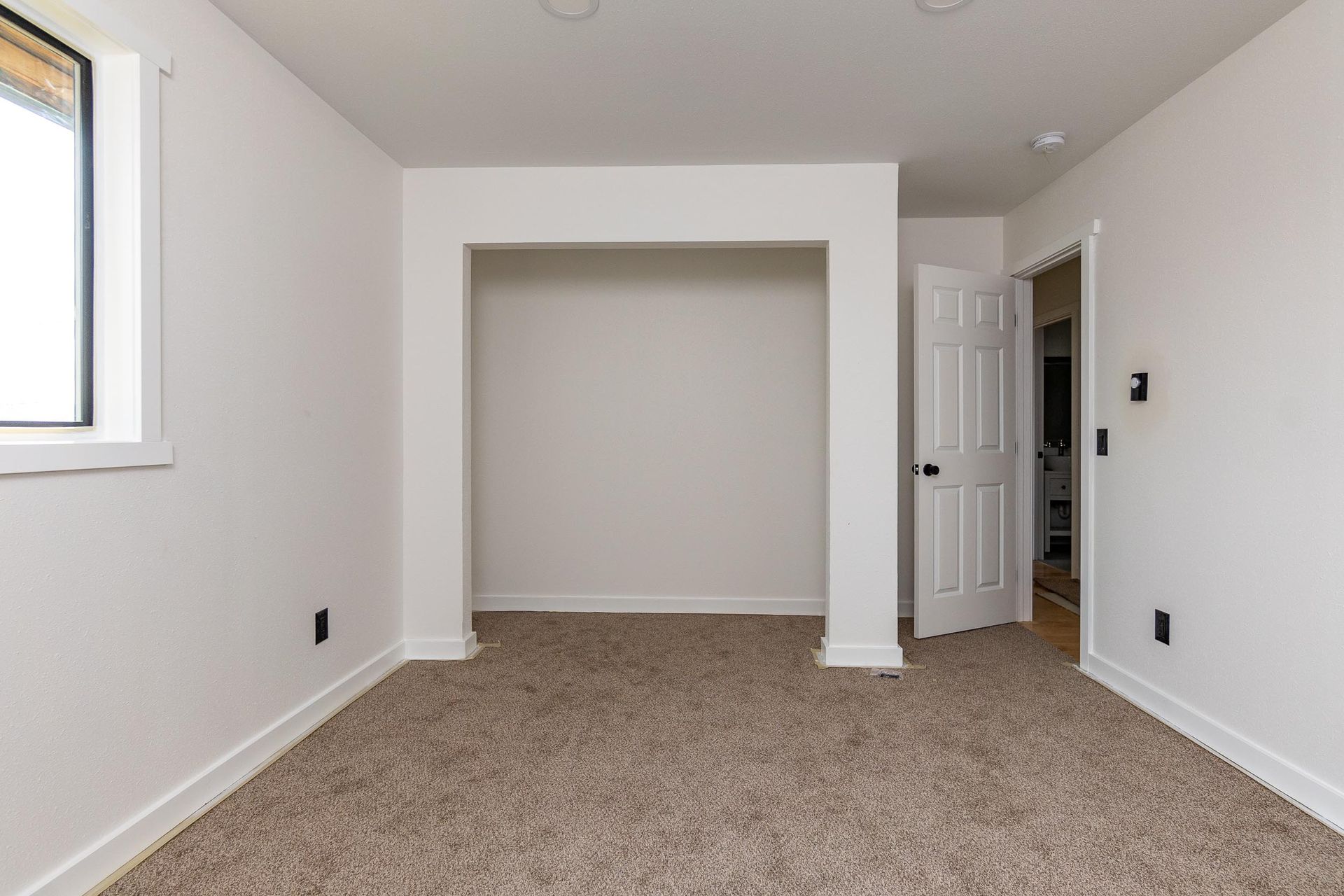 Empty room with beige carpet, white walls, closet alcove, and open door to a bathroom.