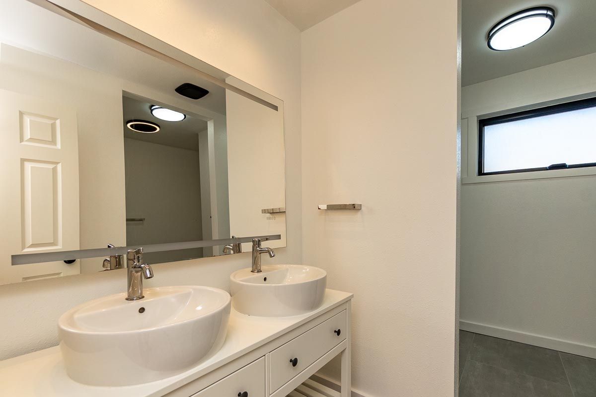 Bathroom with double sinks, large mirror, and open doorway to a separate toilet area. White and gray tones.