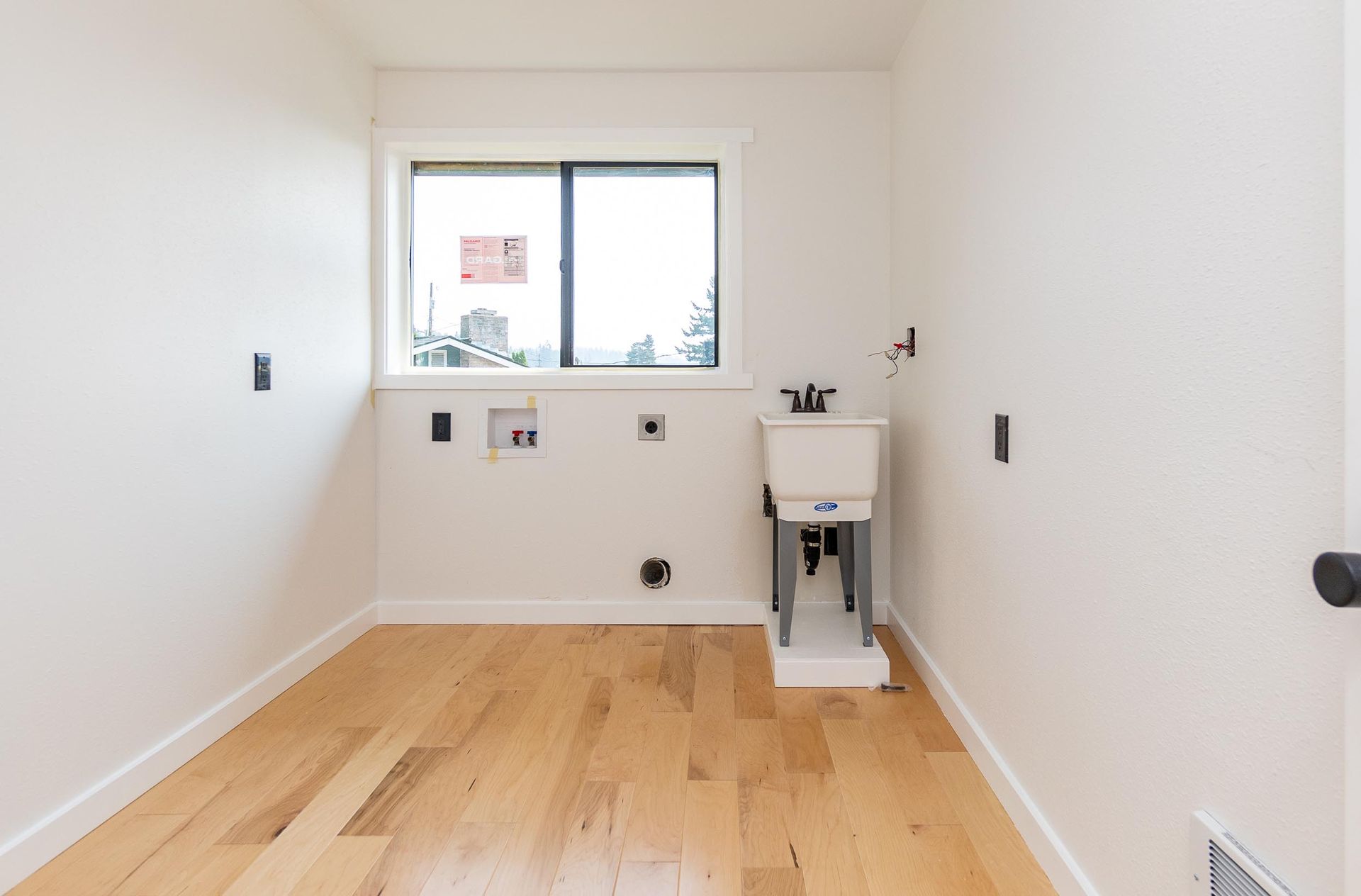Empty laundry room with a window, sink, and light wood floors. White walls and trim.