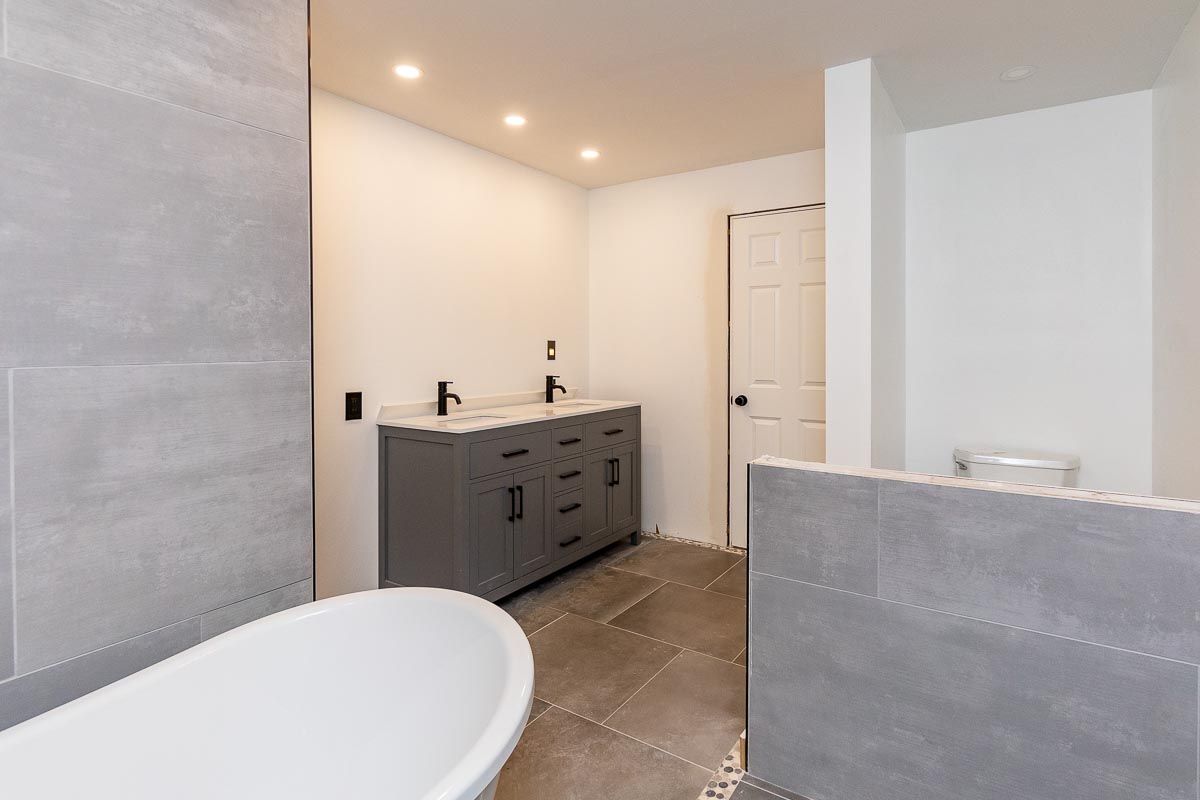 Bathroom with grey vanity, white tub, grey tile walls and floors, and a white door.