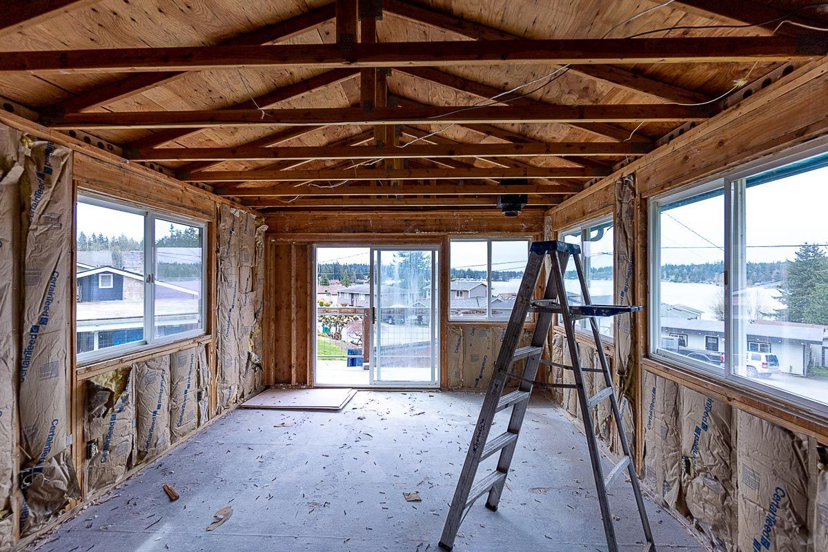 Interior of a room under construction, with exposed wooden beams, insulation, windows, and a ladder.