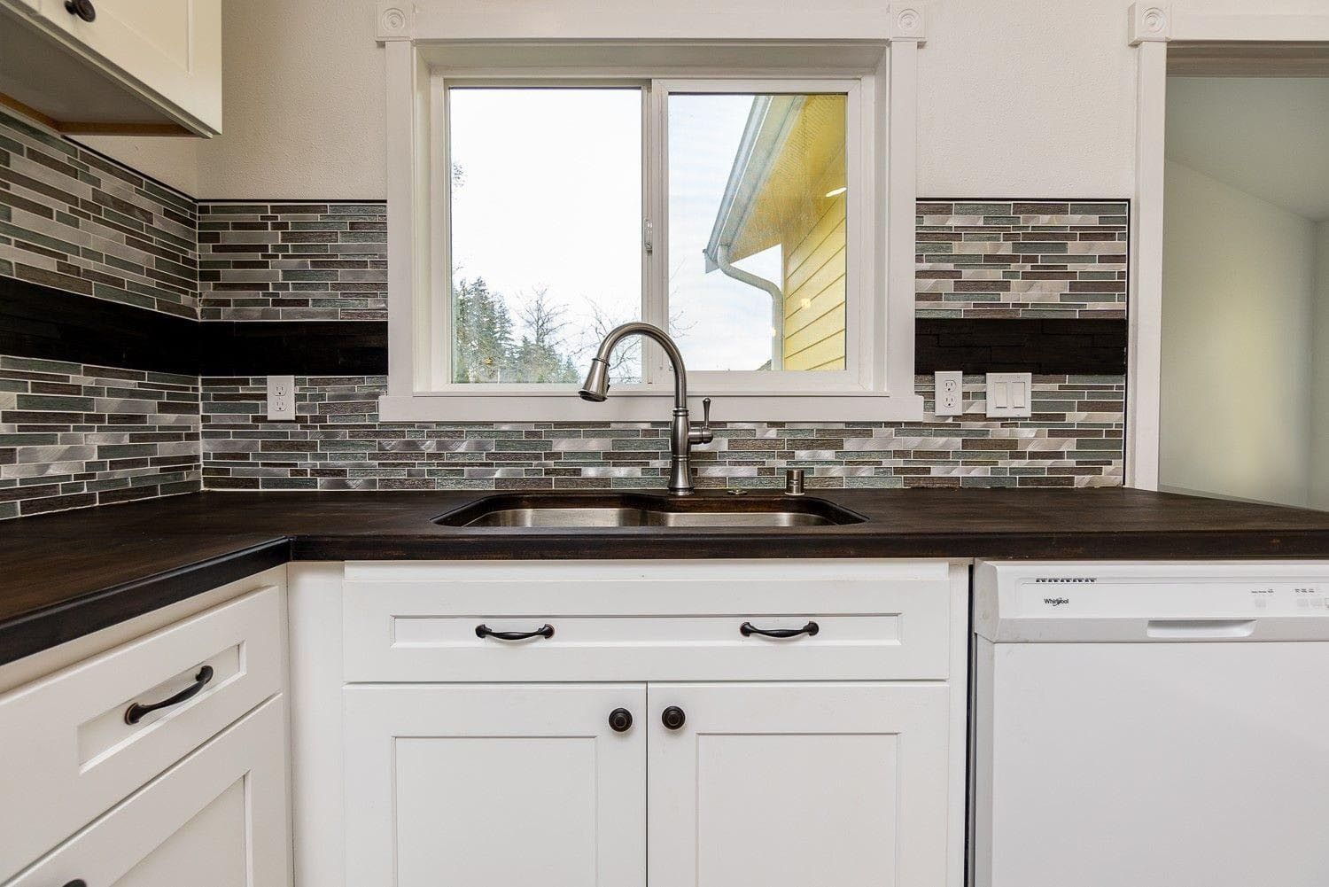 Kitchen with white cabinets, dark countertop, and mosaic tile backsplash; window over sink.