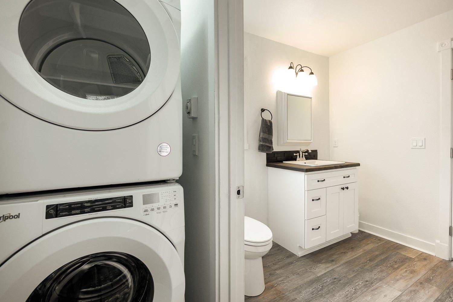 Stacked white washer and dryer next to a bathroom with a white vanity, toilet, and grey flooring.