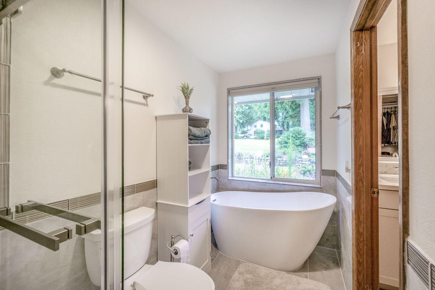 Bathroom with a modern white tub, glass shower, toilet, shelving, and a window overlooking greenery.