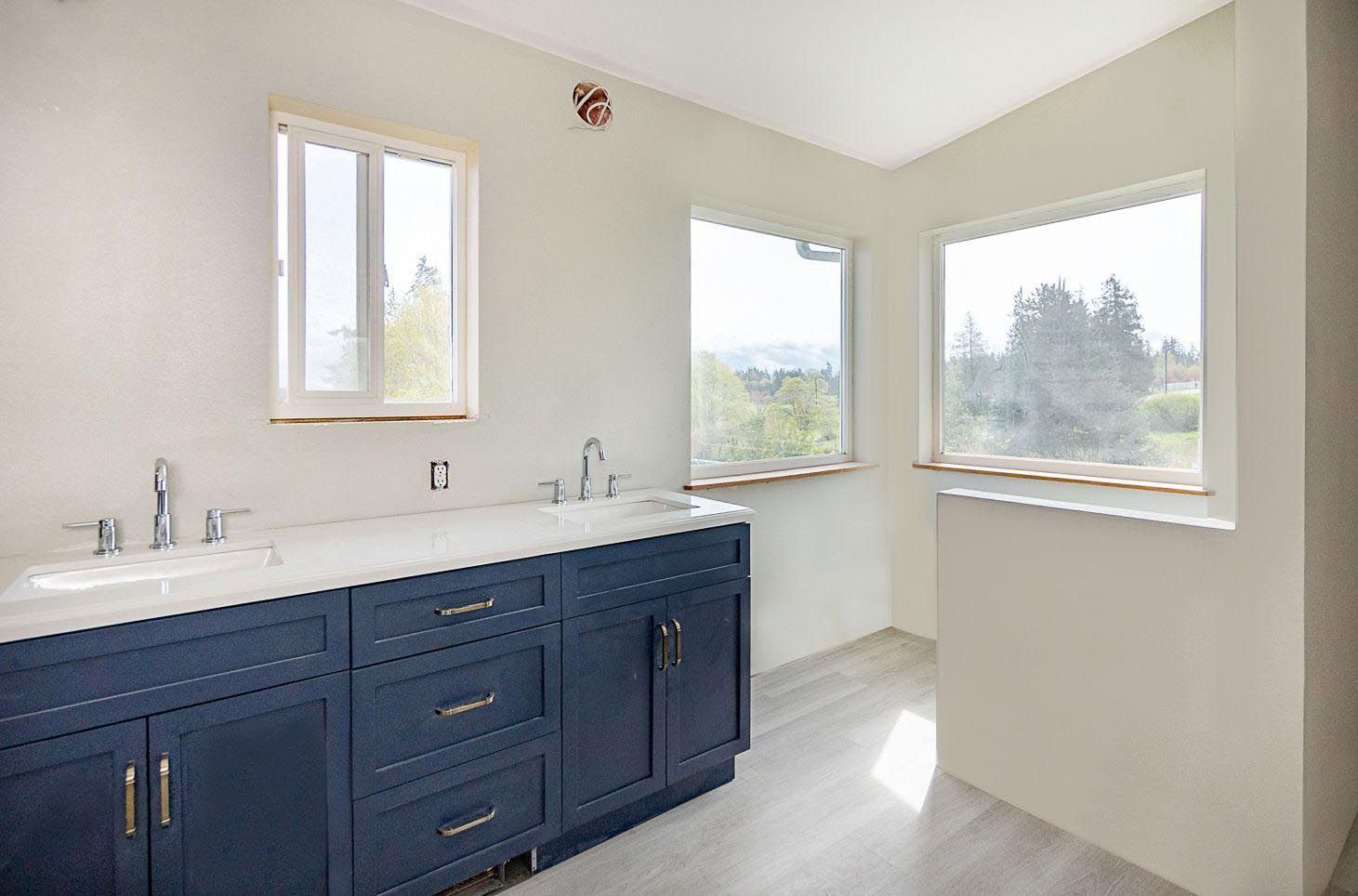 Bathroom with blue cabinets, white countertop, two sinks, three windows, and light gray flooring.