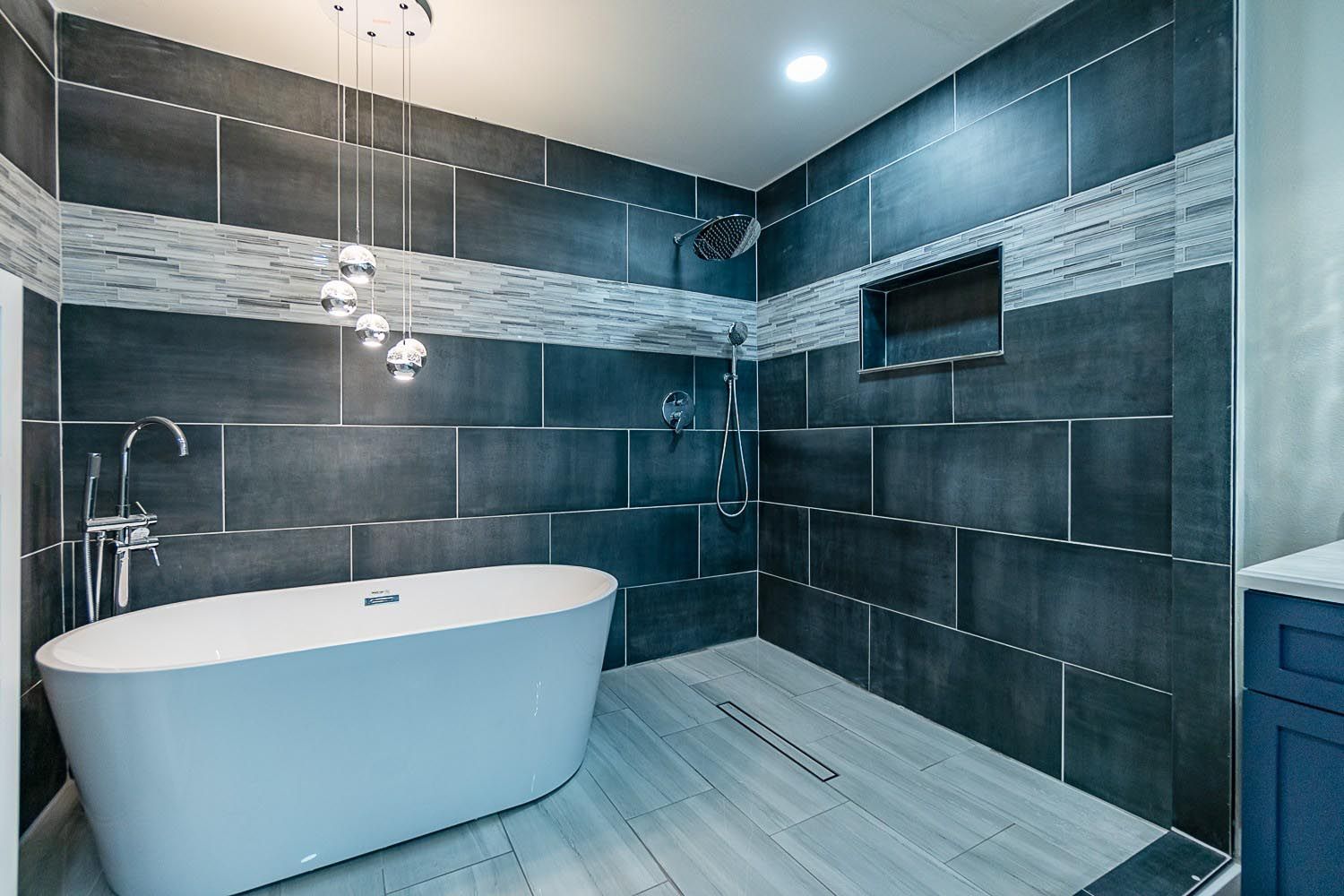 Modern bathroom with gray tiled walls, a white freestanding tub, and overhead lights.