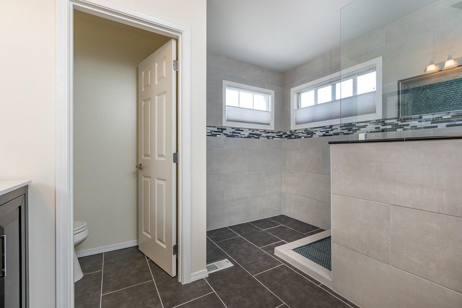 Bathroom interior with a shower, toilet, and open door. Gray and beige tile, two windows with blinds.
