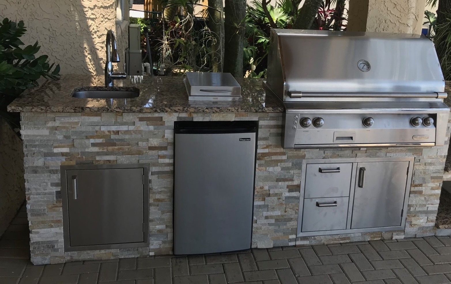 Outdoor kitchen with a grill, sink, refrigerator, and storage cabinets made of stone.