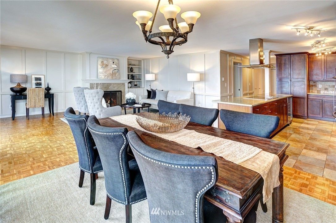 Dining room with a wooden table, blue chairs, and a chandelier. A kitchen is in the background.