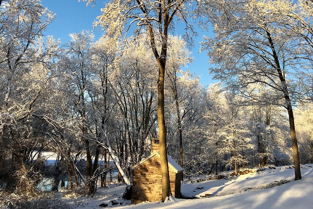 Snow-covered trees frame a stone structure and a body of water under a bright blue sky. Sunlight bathes the scene.