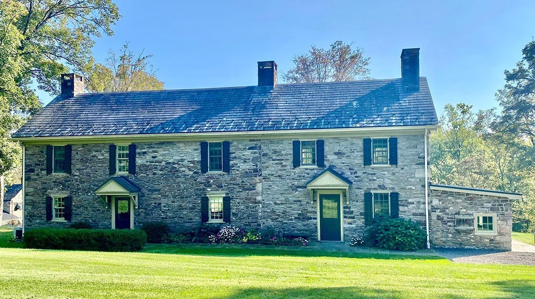 Two-story stone house with dark shutters, three chimneys, and a slate roof, set on a grassy lawn.