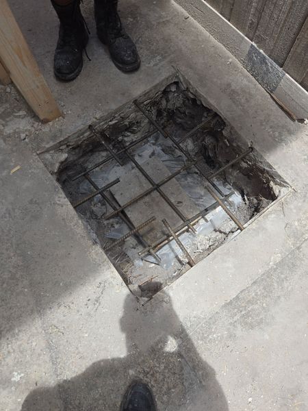 Open square hole in concrete floor with exposed rebar, person's legs and shoes visible nearby.