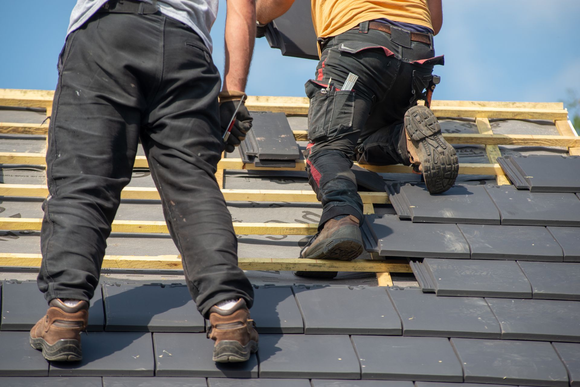 Two roofers installing dark gray roof tiles on a wooden frame under a blue sky.