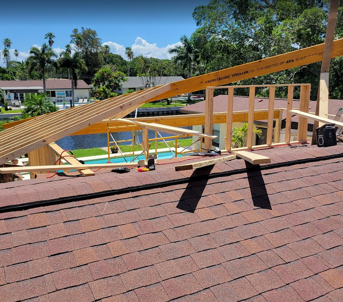 Construction of a wooden structure on a brown shingle roof with a pool and waterfront in the background.