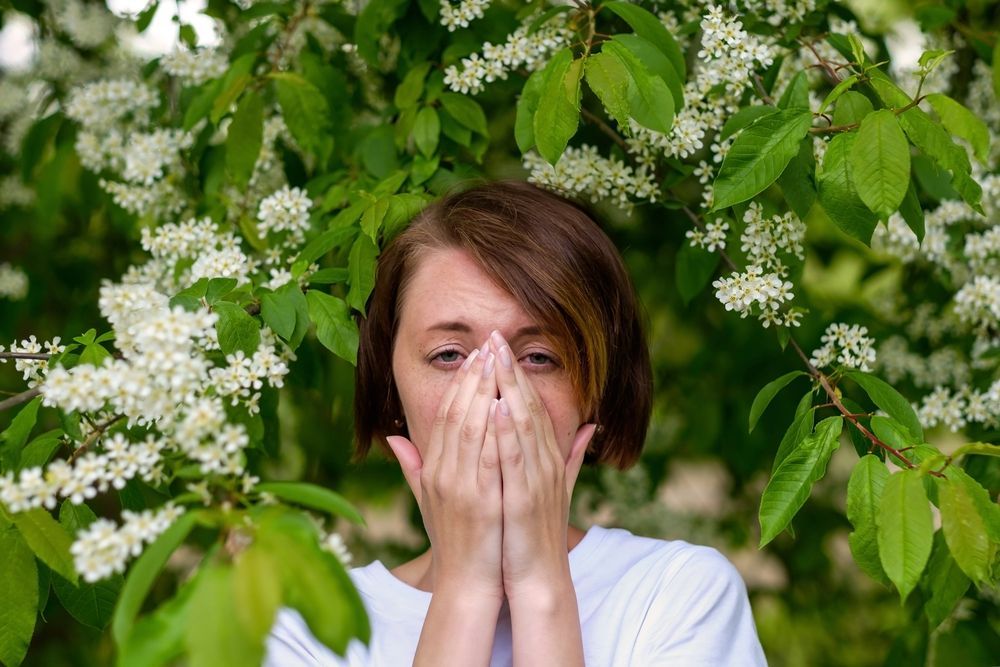 Una persona si copre naso e bocca tra fiori bianchi in fiore e foglie verdi all'aperto