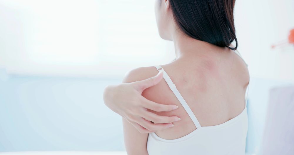 Una donna si tocca la scapola, mostrando segni di mal di schiena in una stanza luminosa.