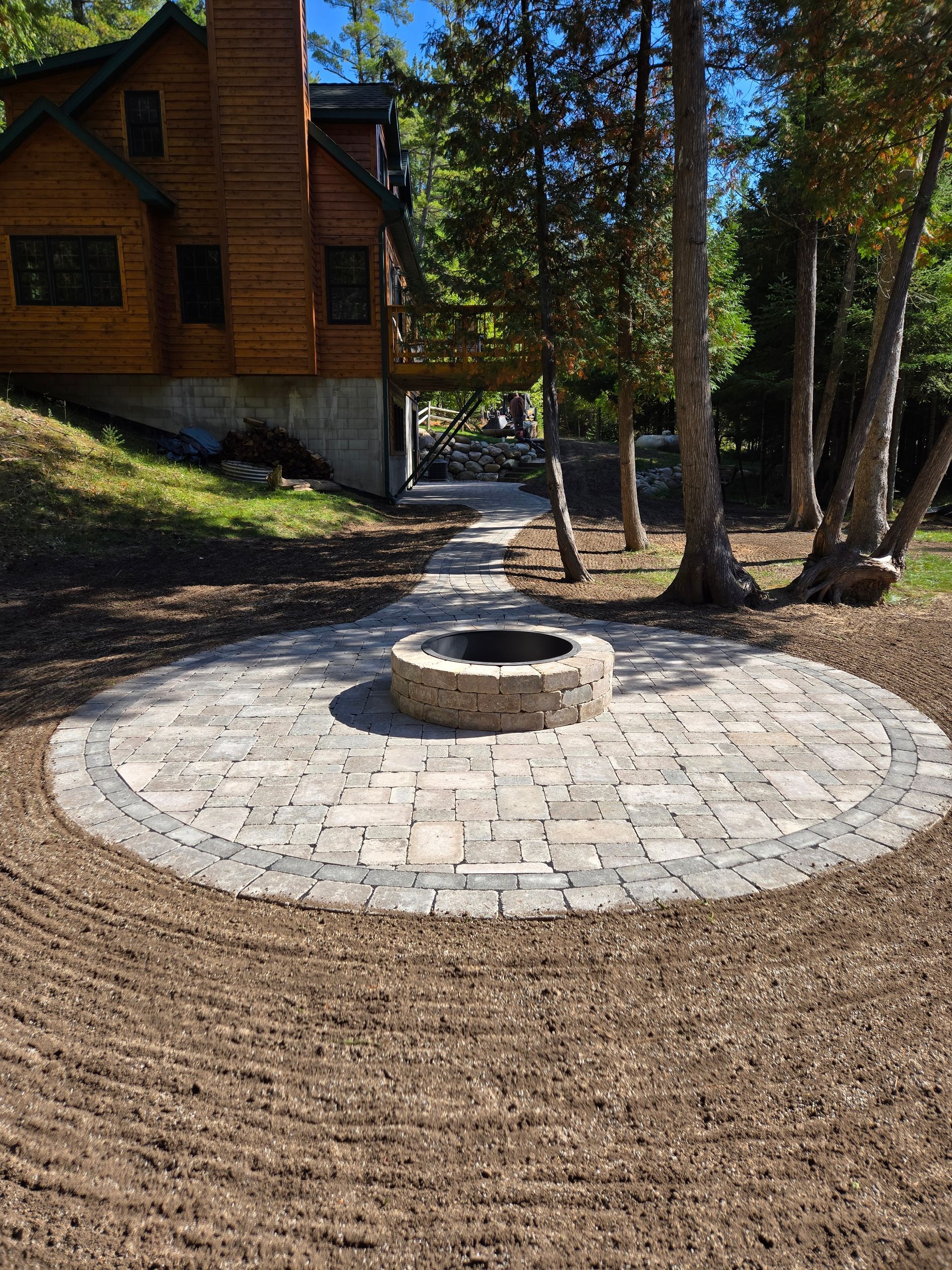 Two workers in a sunny, wooded outdoor setting install a grey, patterned stone walkway. Two workers in a sunny, wooded outdoor setting install a grey, patterned stone walkway.