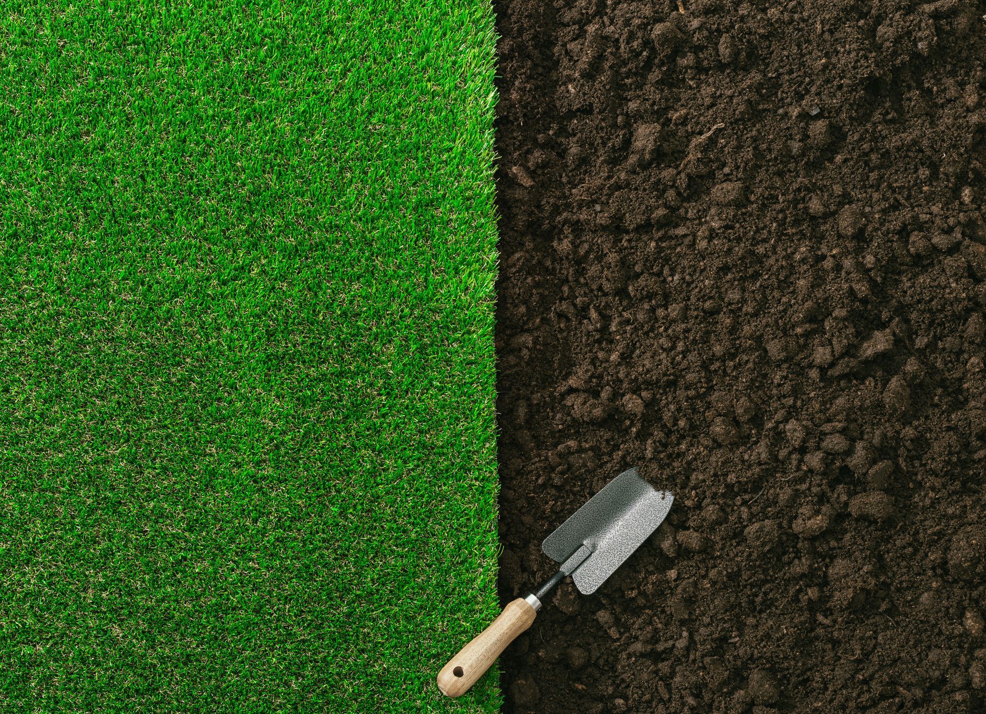 A garden trowel rests on dark soil beside a patch of vibrant green artificial grass, split vertically down the middle.