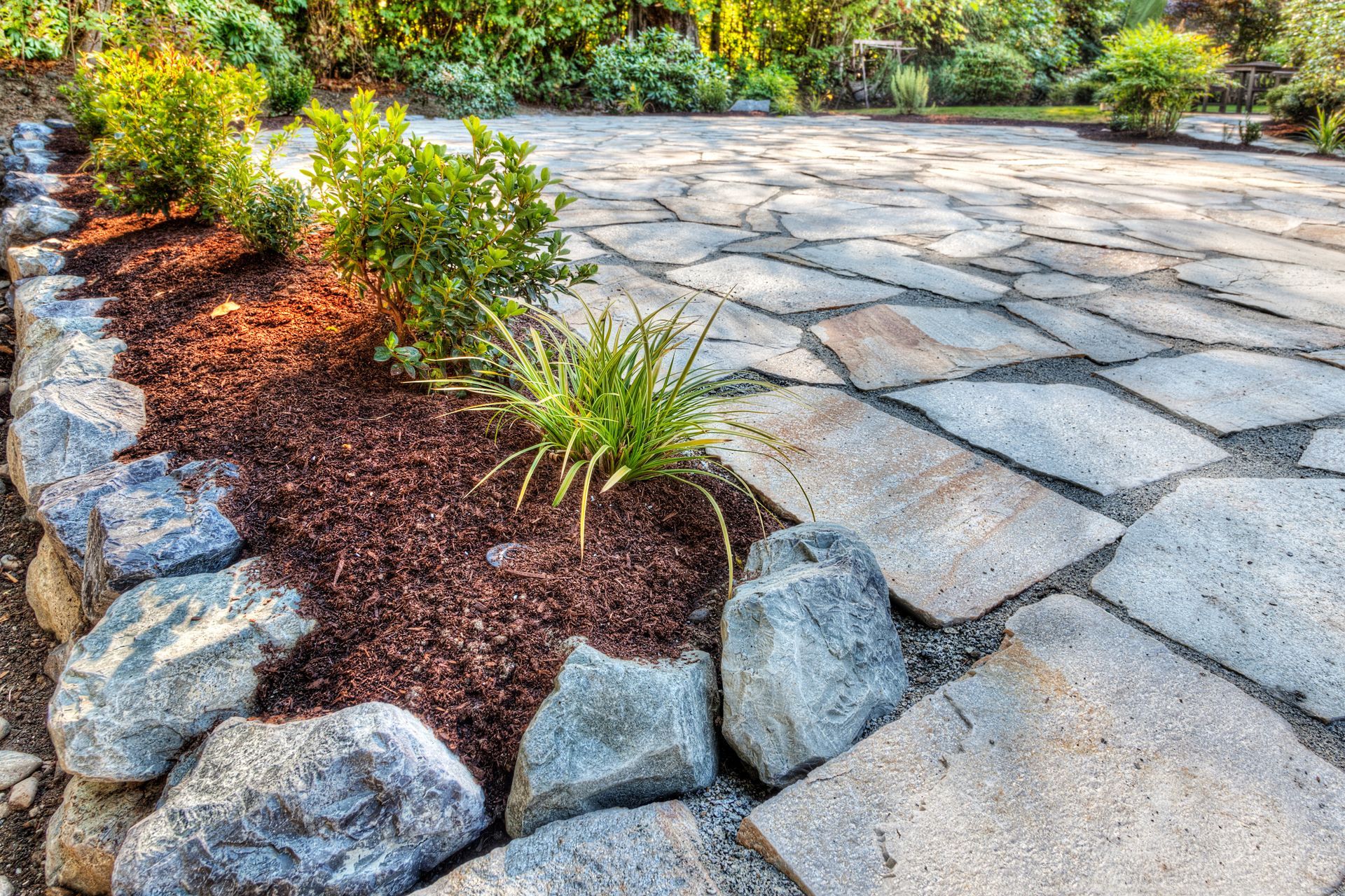 A stone patio borders a garden bed filled with dark mulch, small green shrubs, and decorative gray boulders.